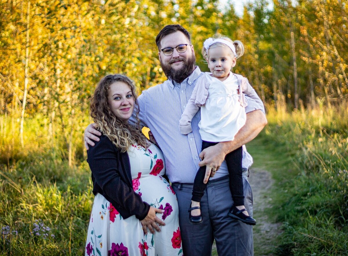 A woman in a floral dress holds her pregnant belly next to her husband who is holding their toddler daughter