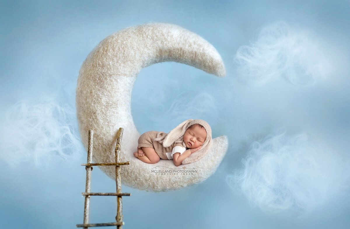 Newborn baby sleeping on a blue crescent moon prop with fluffy clouds in a studio setup