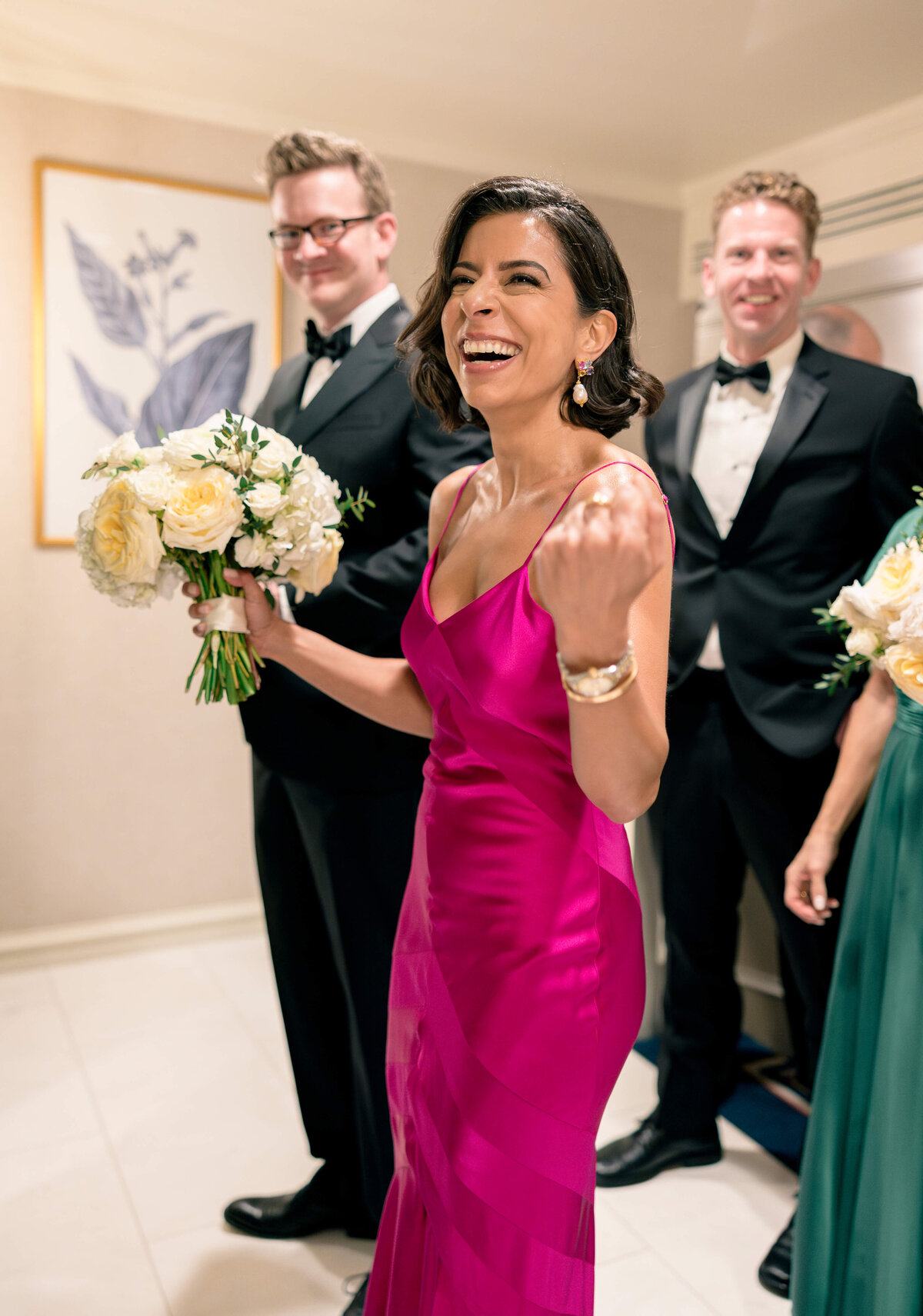 A woman in a vibrant pink dress smiles joyfully while holding a bouquet of white flowers. She is surrounded by two men in tuxedos and another person in a green dress. They appear to be in a celebratory setting, possibly a wedding.