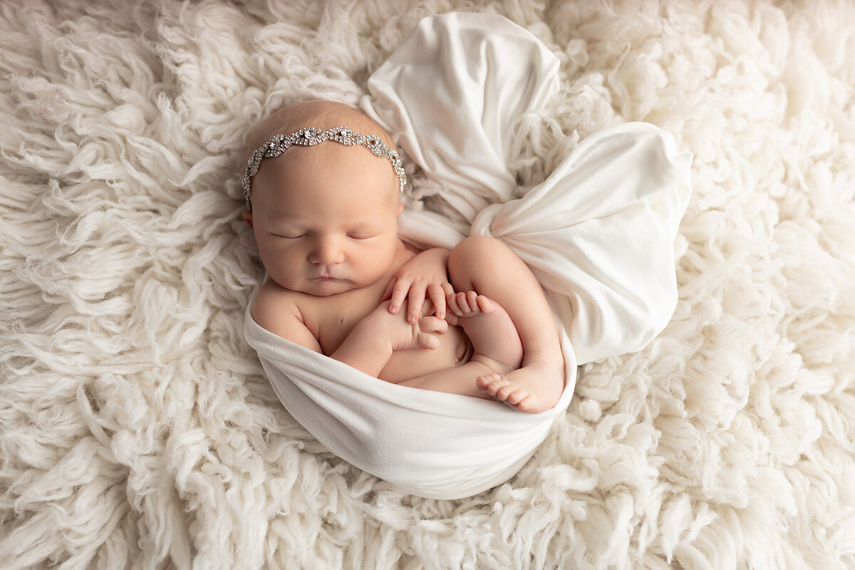Beautiful newborn baby girl wrapped in white with rhinestone headband, captured by Dallas Newborn Photographer specializing in timeless newborn pictures.