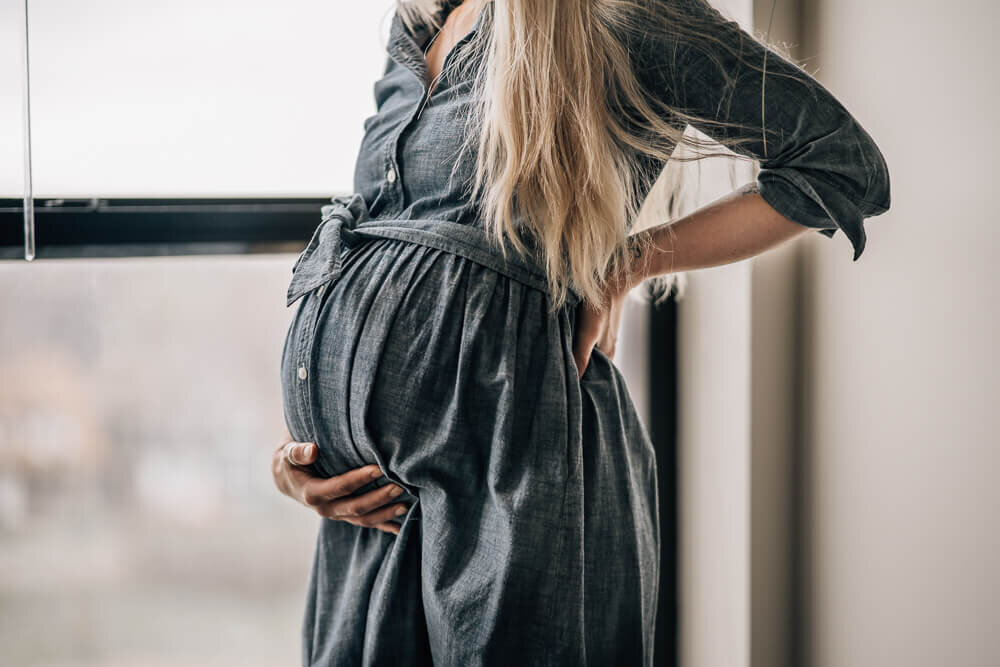 An expecting mama stands angled in front of her window in a denim dress during her in home maternity photography session by Kate Simpson.