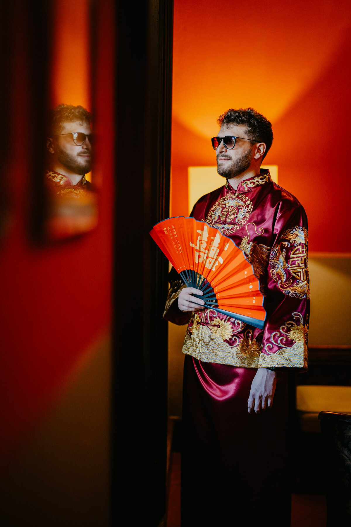 Groom in Chinese red outfit with sunglasses and fan, wedding photographer Tuscany.