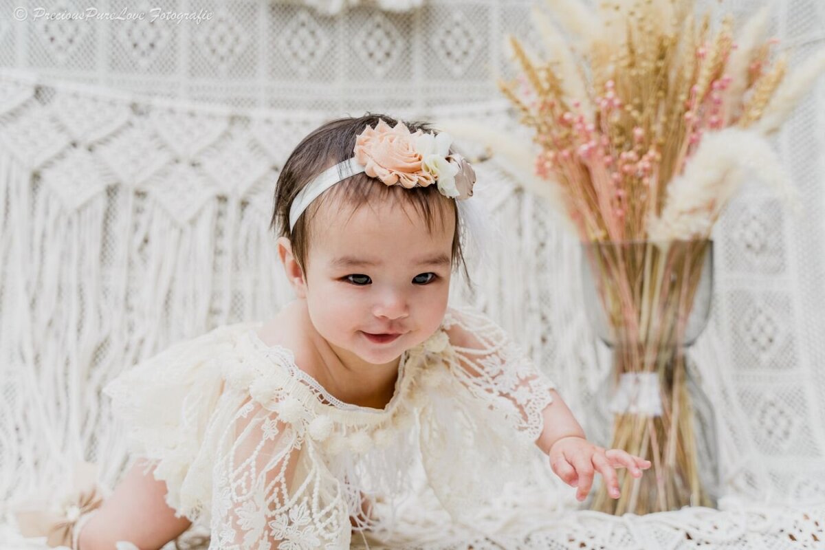 Baby girl in an ivory lace romper and pastel floral headband crawling on a cream crochet mat with a vase of pampas grass behind her.