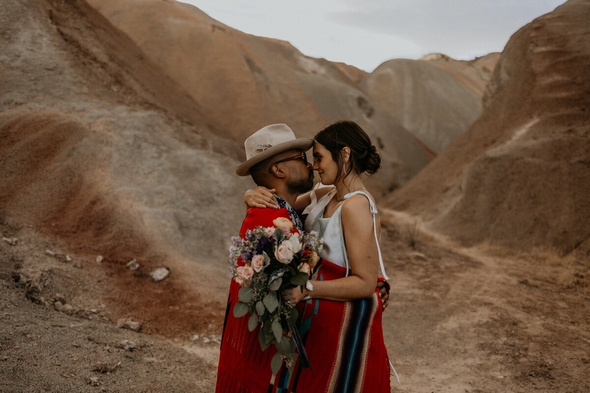a groom wrapped in a native American blanket hugging his new bride in the desert