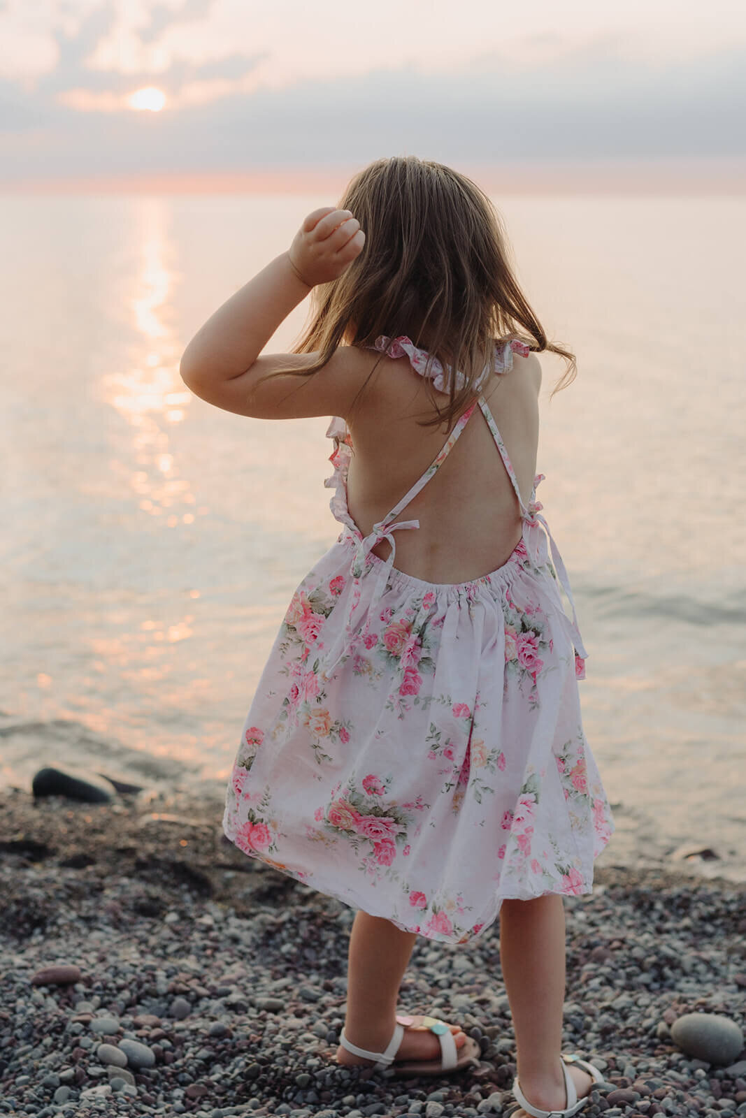 Little girl in floral dress standing by waters edge at family with two kids on the beach at family photo shoot in Finger Lakes, NY