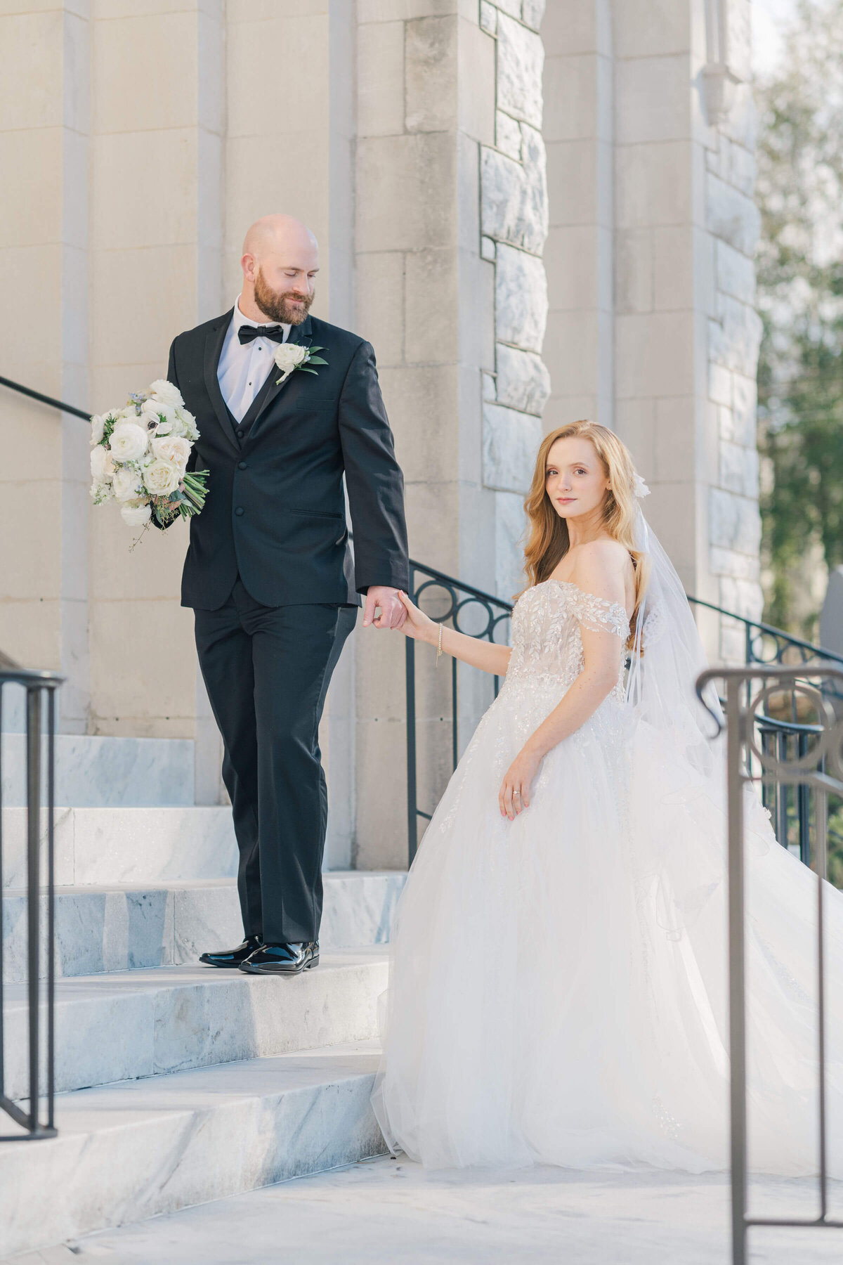 Bride and groom on stairs with groom standing abvoe bride holding her bouquet and brides hand while she looks at the camera