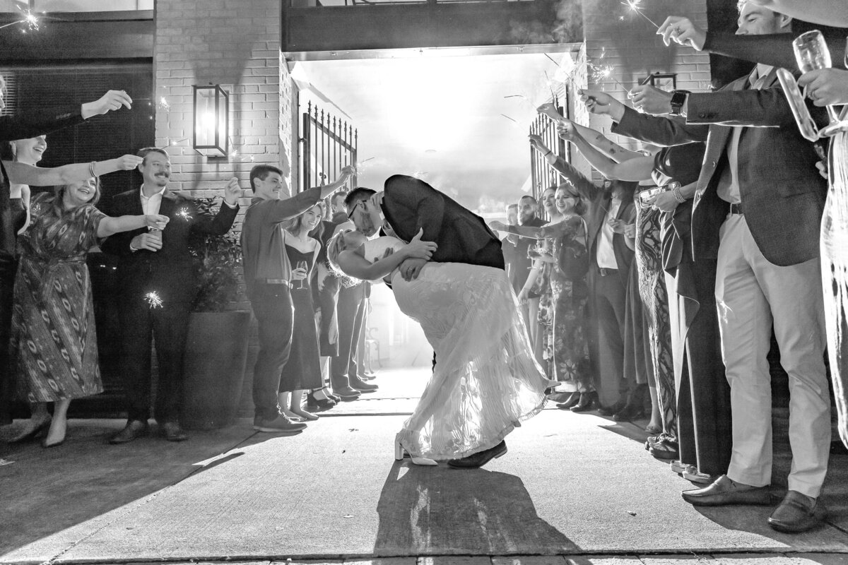 A bride and groom share a kiss as the groom dips the bride, surrounded by cheering guests holding sparklers and celebrating their wedding at a nighttime outdoor exit.