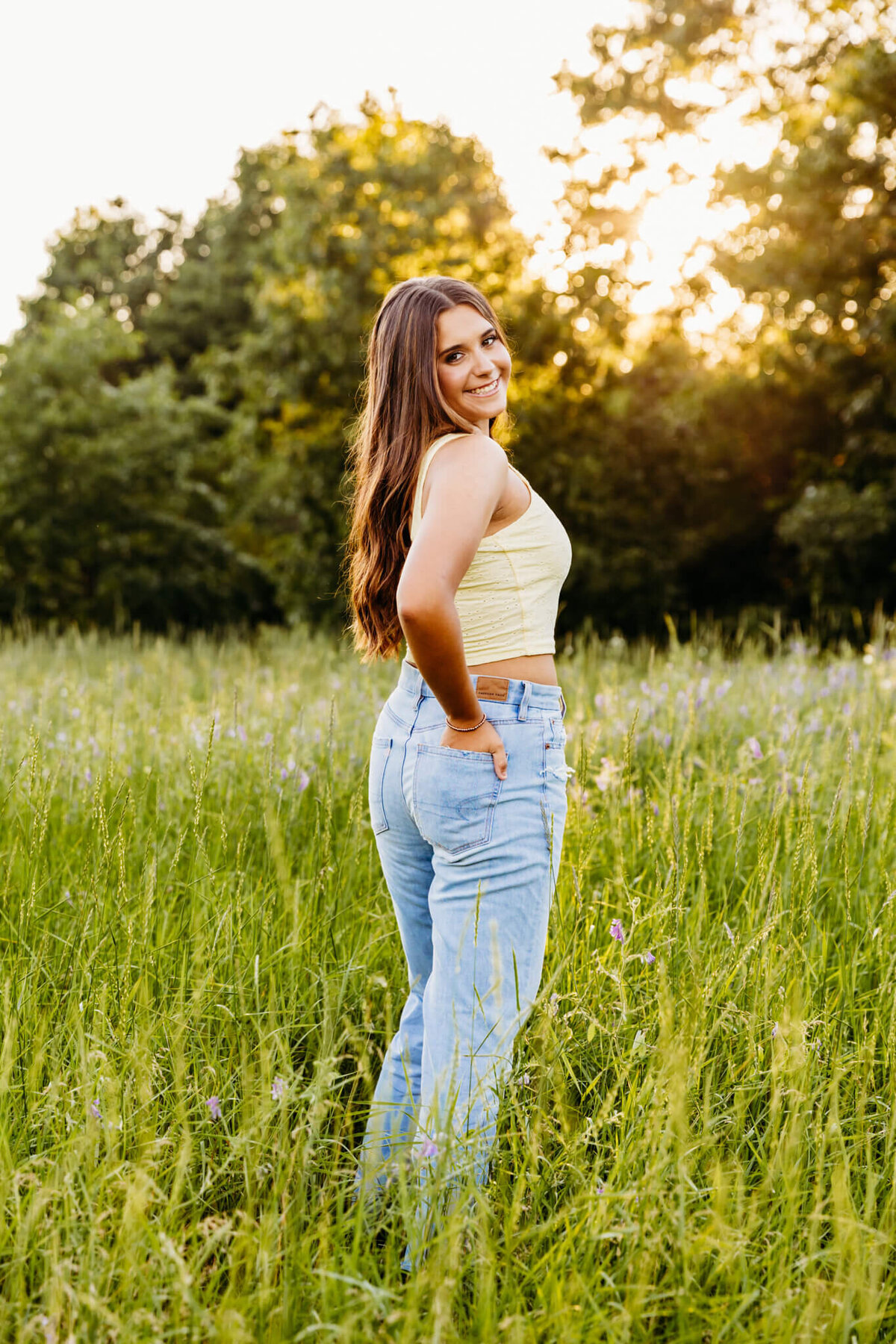 curly brown haired girl standing in a glowing field with her hand in her back pocket as she looks over her shoulder for her senior photos