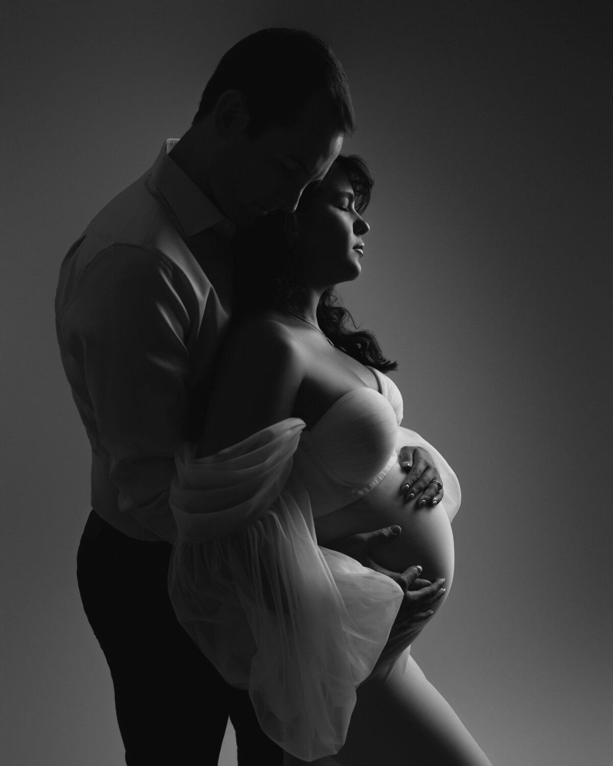 Black and white romantic studio portrait of couple holding baby bump with soft directional lighting.