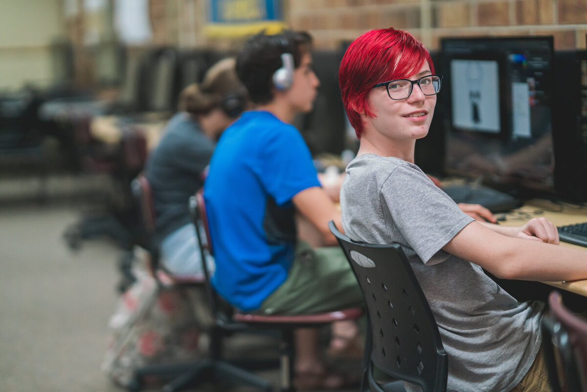 Student with hot pink  hair smiling at the camera while sitting in a computer lab at school
