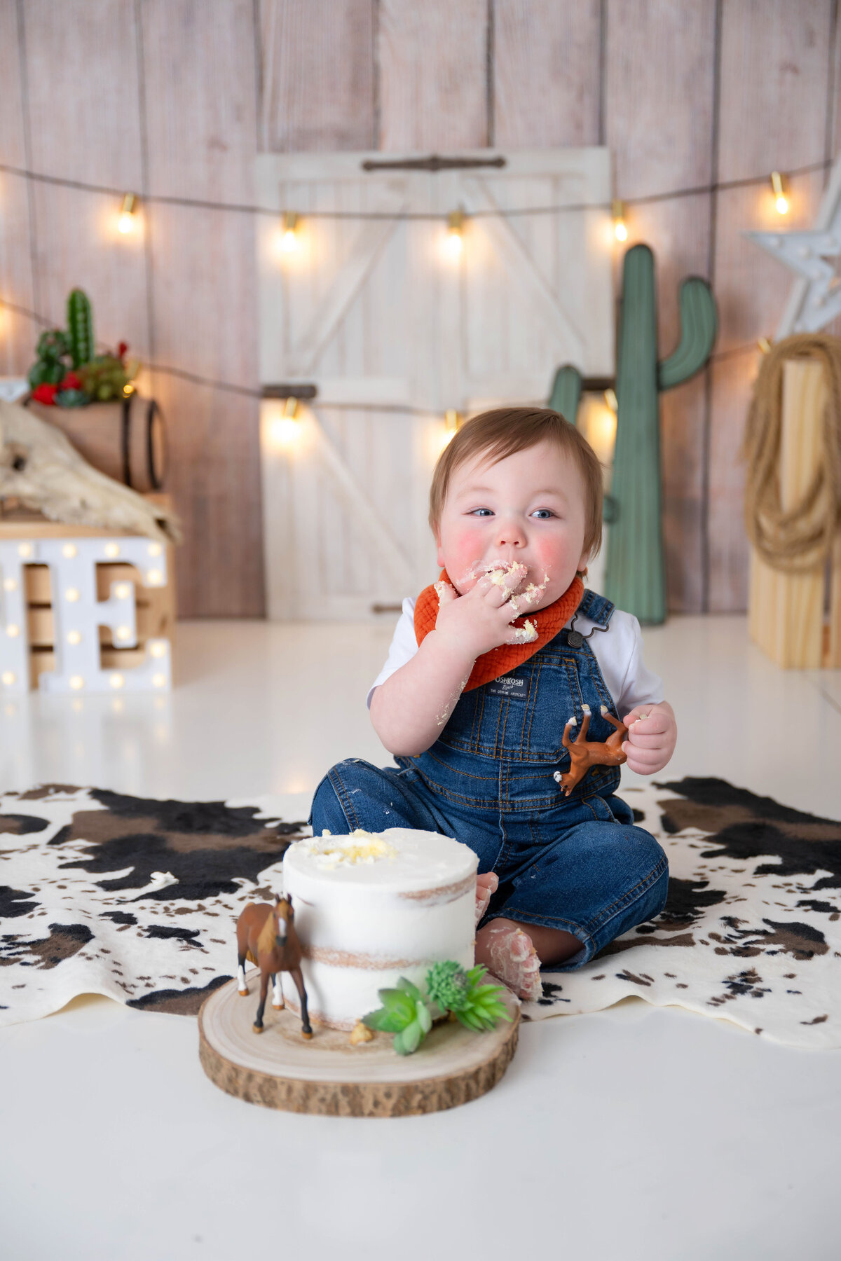 Baby smiling, looking right, licking cake off fingers with cowboy props nearby.