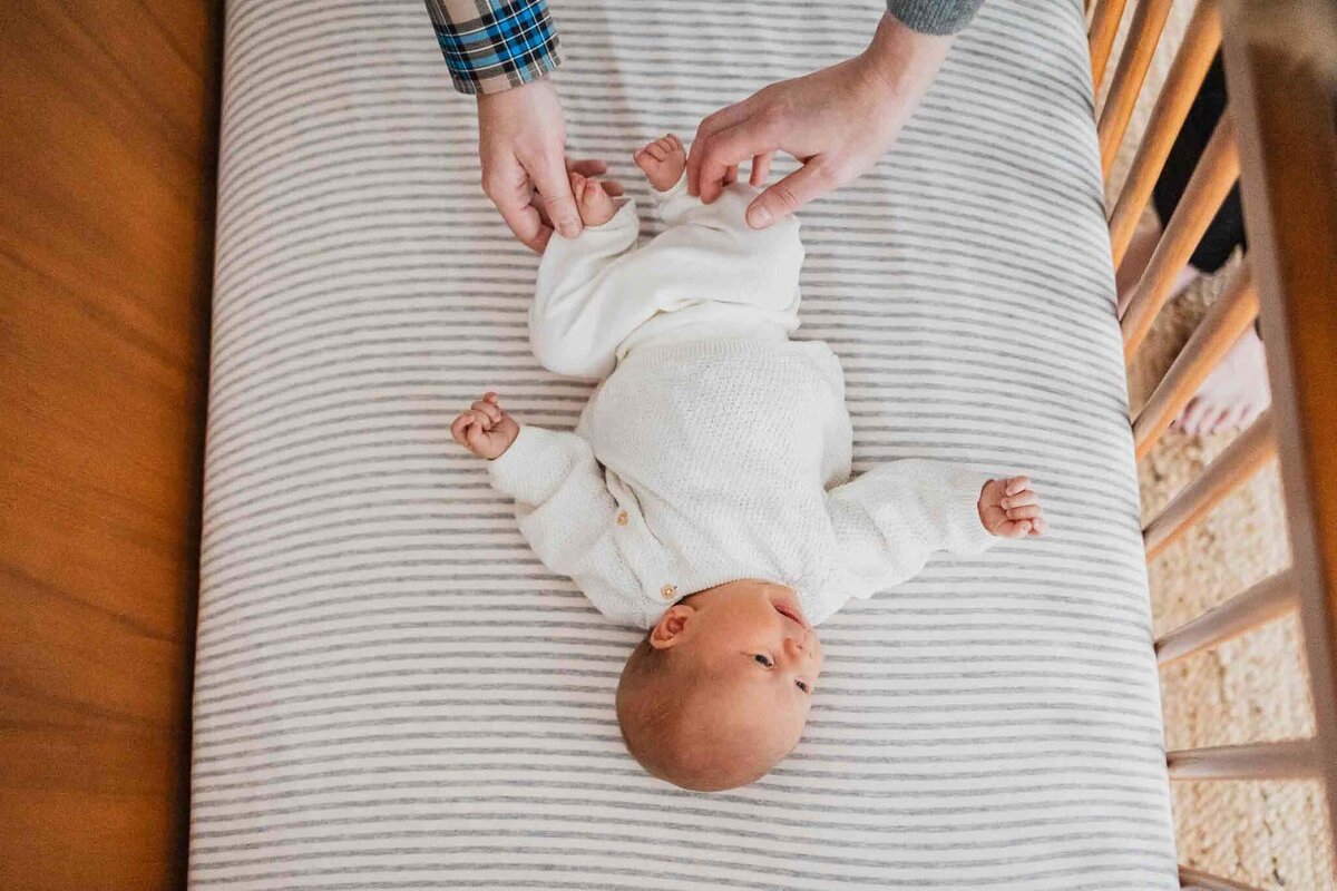 A cozy newborn lying in a crib with striped bedding, lovingly held by hands adjusting their outfit. Taken in a serene Cleveland nursery setting.