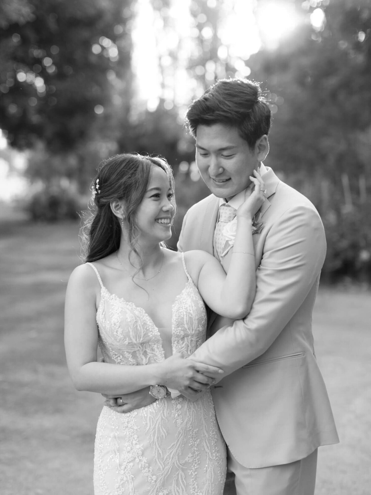 A joyous couple embraces outdoors in a black-and-white photo. The bride gazes at the smiling groom in a light suit.