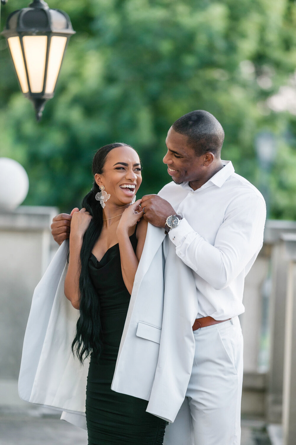 Couple at Adriatica Village during their engagement session, she laughs as he drapes his jacket over her shoulders while they look at each other.