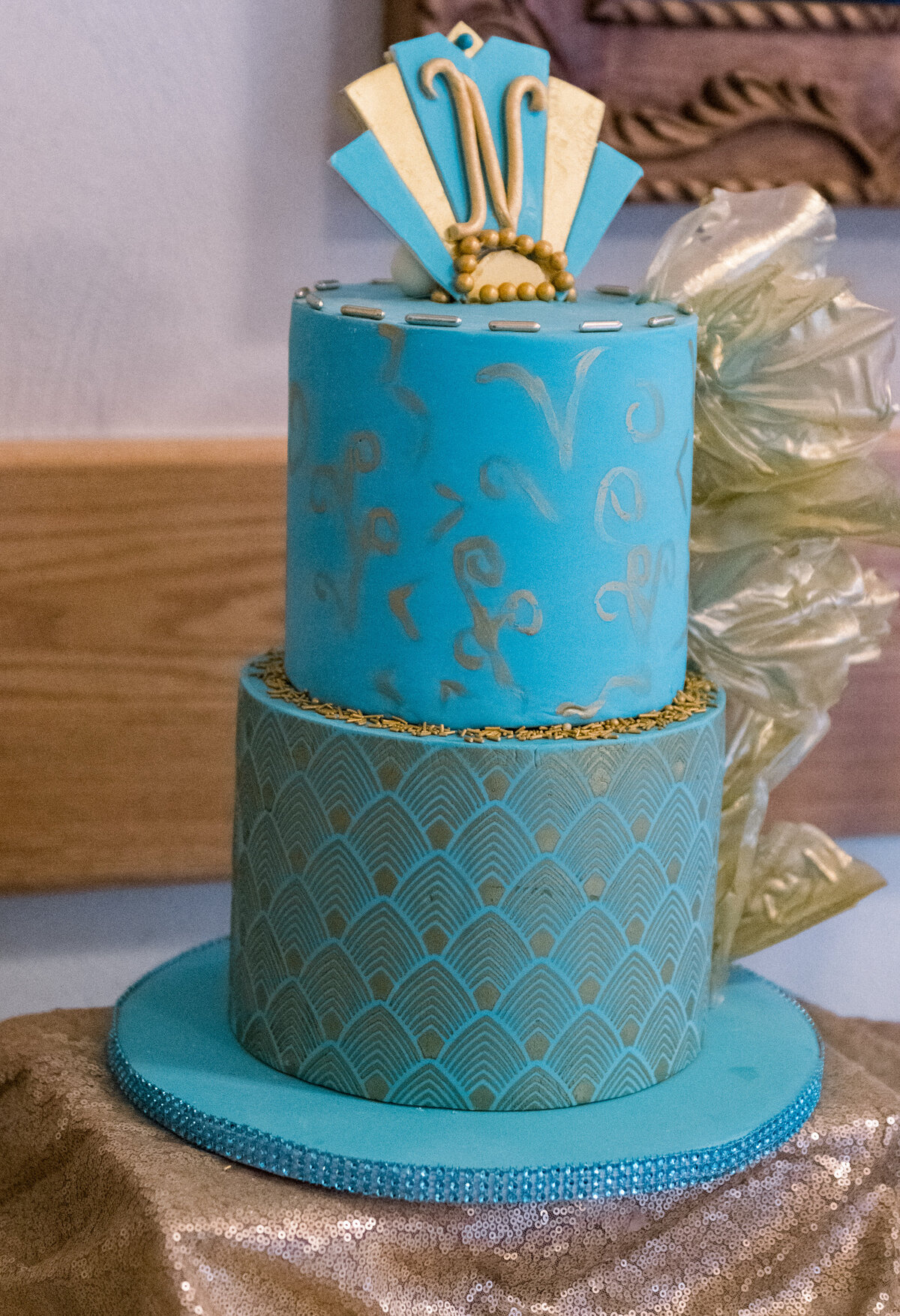Art deco design 2 tier cake. in teal and gold. Art deco fan stencil on bottom tier; handpainted abstract lines on top tier; art deco fan monogram  cake topper. Rice paper gold sails on side edge of cake