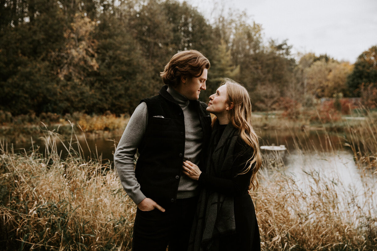 Man and woman standing in front of pond and walking trails for fall engagement photo. The man's body is facing the camera, the woman is facing the man. They are standing close, looking at each other, and smiling.
