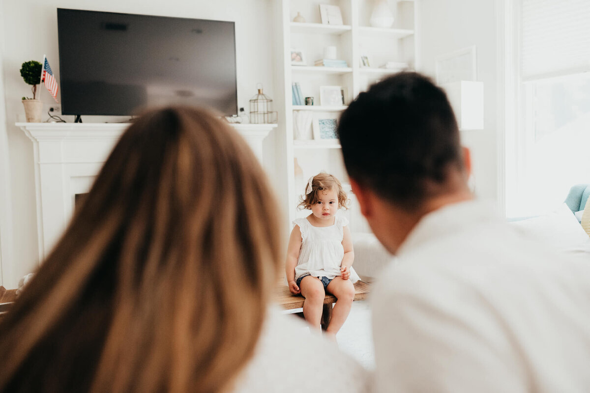 A mother and father look at their toddler daughter as she sits on the coffee table in a cute white outfit in their living room