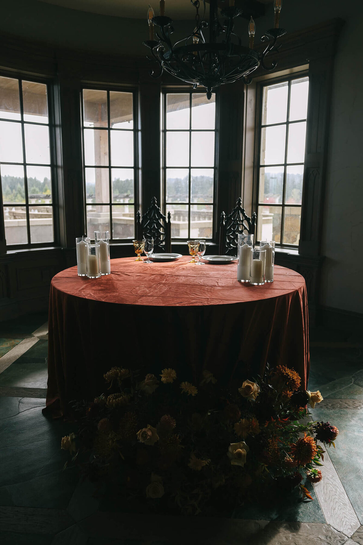 A table draped with a table cloth and set with candles and tableware with flower arrangements on the floor in front of it.