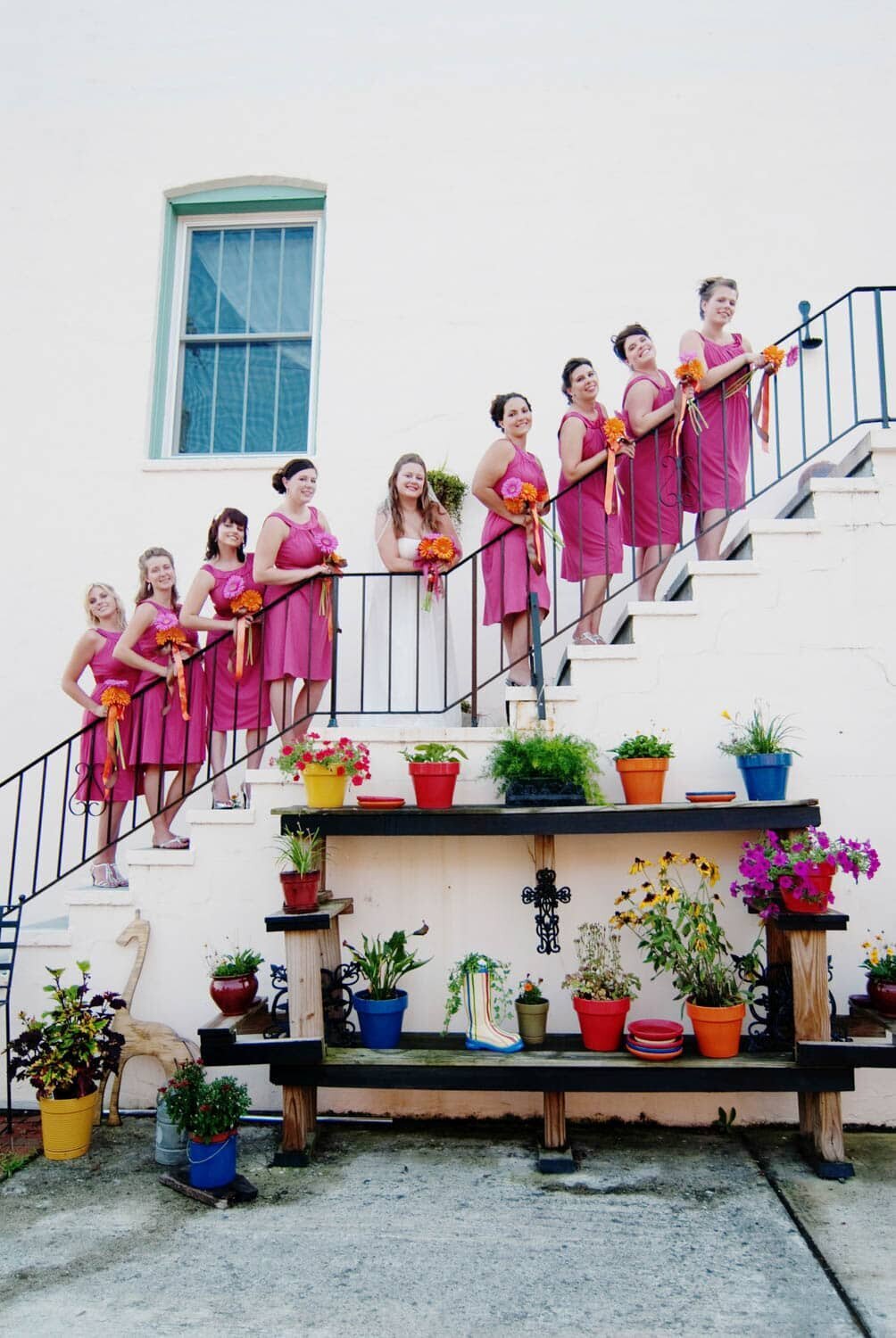 bridesmaids in pink dresses stand on a stairway overlooking a colorful wall of potted flowers