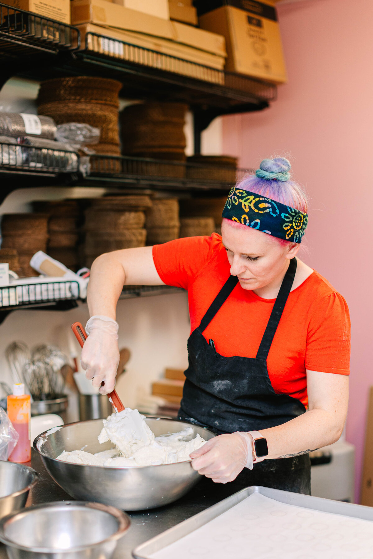 A baker wearing a bright red shirt, black apron, and a colorful headband mixes a large bowl of batter in a commercial kitchen. Shelves stocked with baking supplies and ingredients are visible in the backgroun