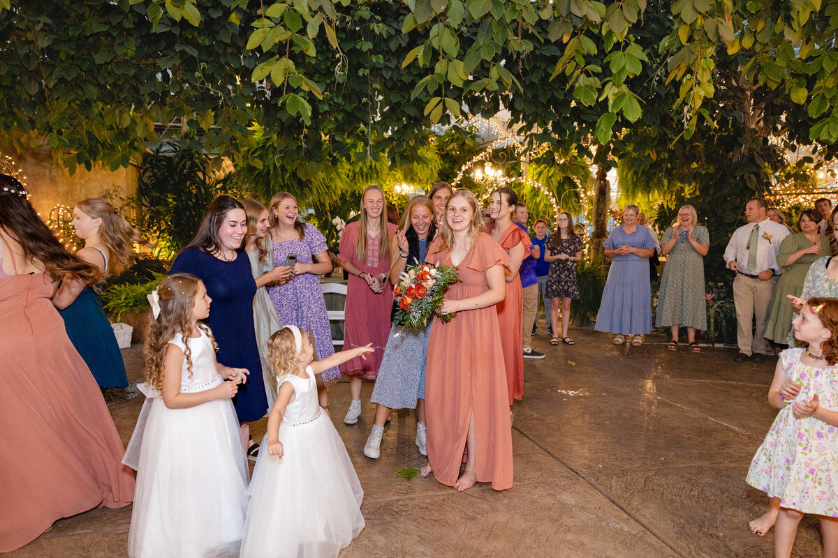A bridesmaid in a flowing rust-colored dress holds the wedding bouquet, surrounded by smiling guests celebrating the bouquet toss.