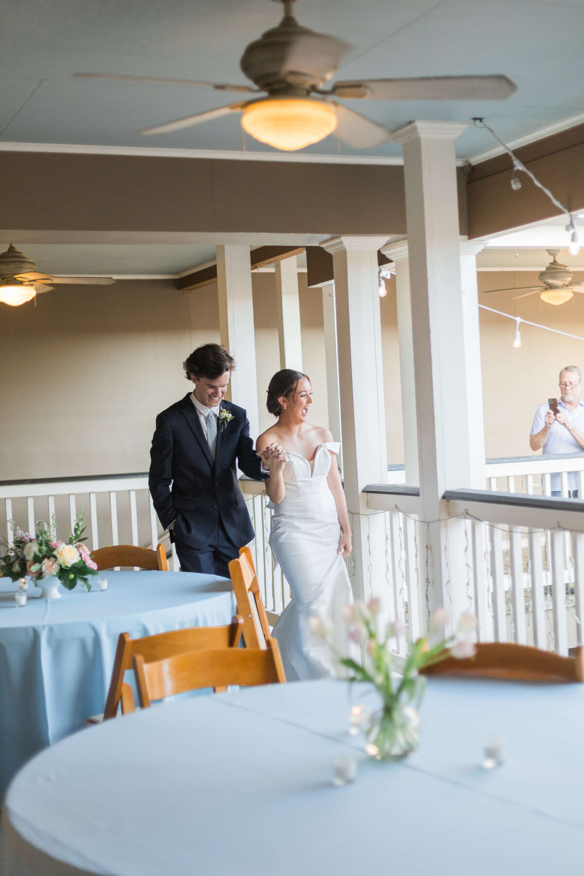 A bride in a white gown and a groom in a dark suit walk together down a balcony with round tables, wooden chairs, and floral centerpieces.