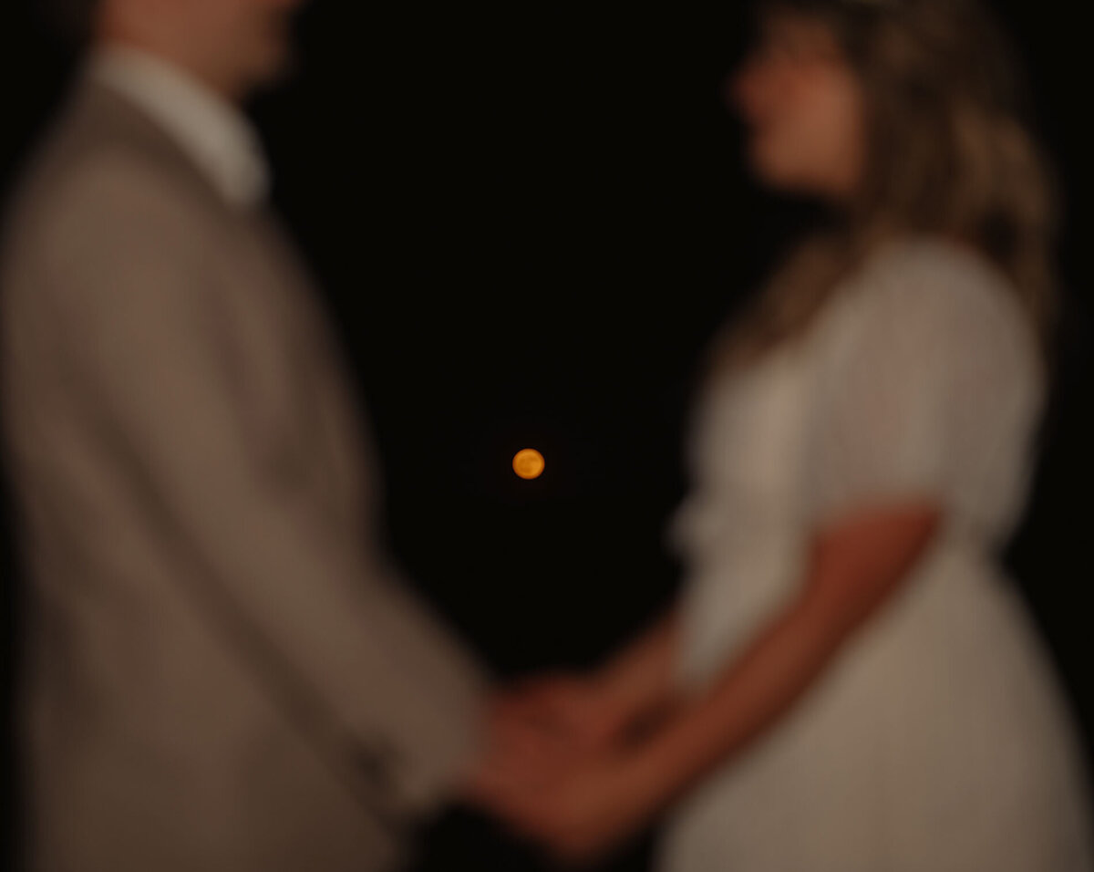 bride and groom hold hands in the foreground while the focus is on the orange, glowing full moon in the background at an ceremony in Bend, OR