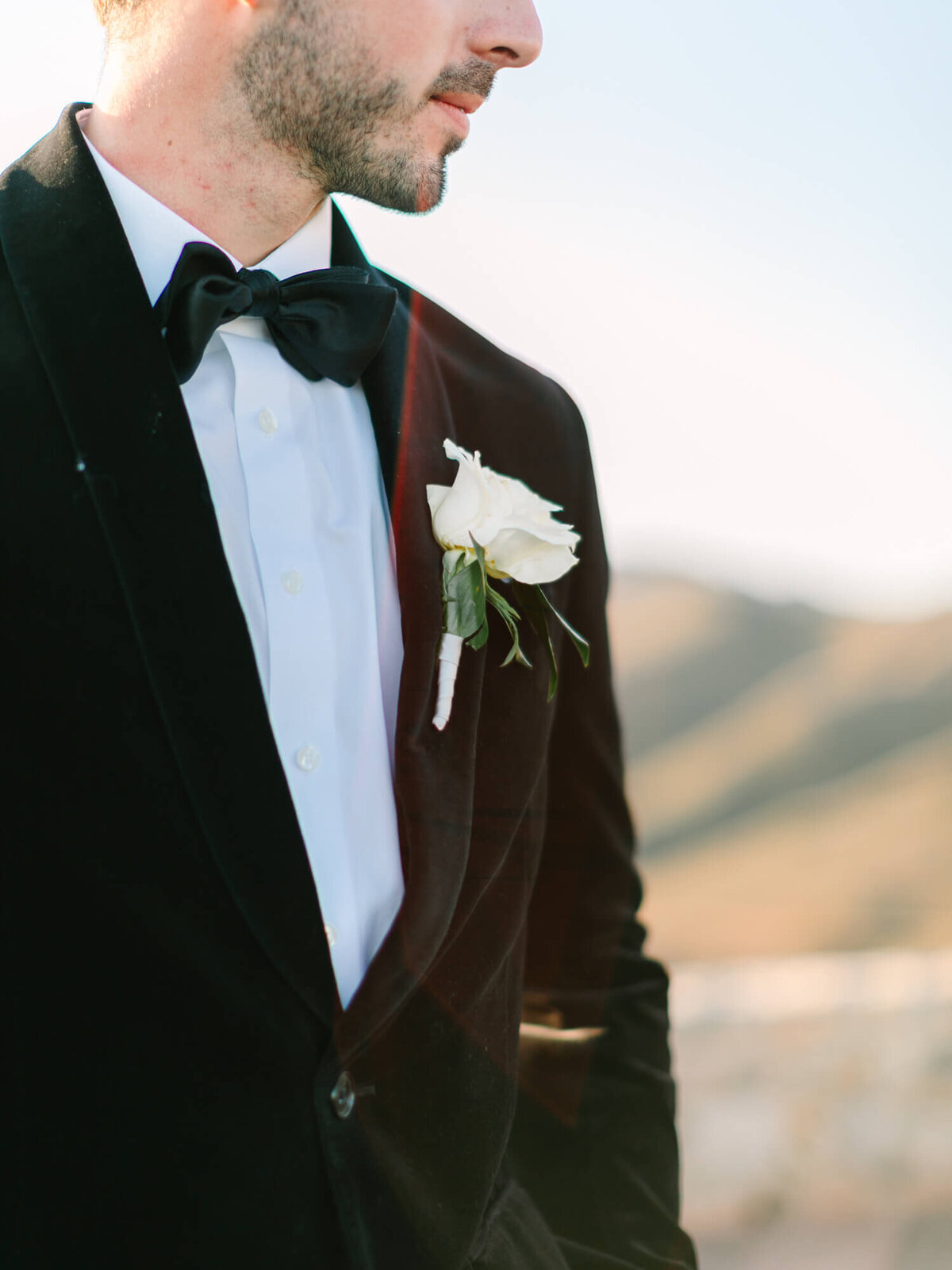 Groom in a black tuxedo with a bow tie and a white rose boutonniere stands outside, with blurred hills in the background