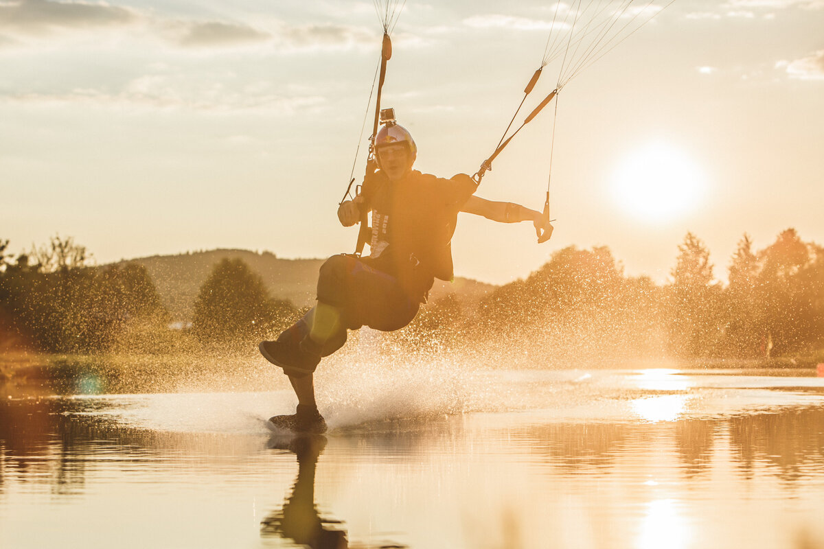 Ein Fallschirmspringer fliegt über das Wasser. Das Bild zeigt einen Fallschirmspringer, der mit einem Fuß im  Wasser streift. Zu sehen ist seine Reflektion und die Umgebung