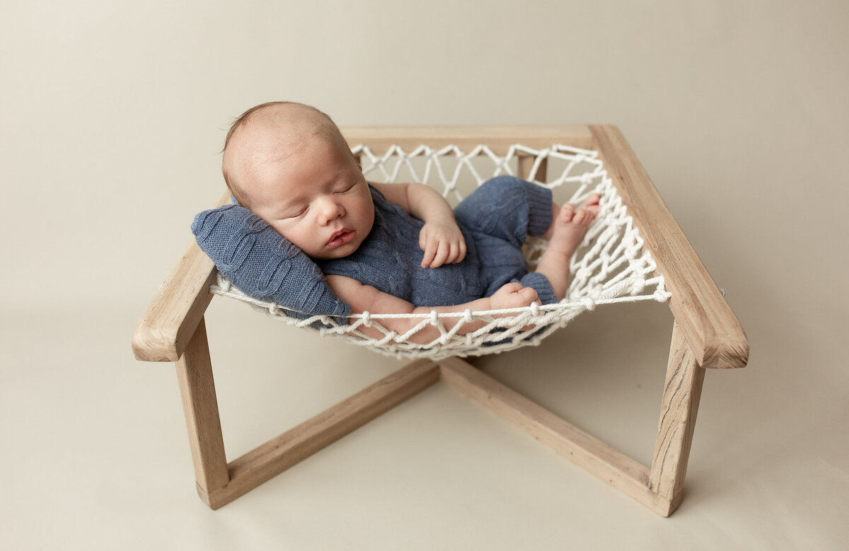 Newborn baby boy laying in a hammock style photo prop in a blue romper, sleeping on a pillow