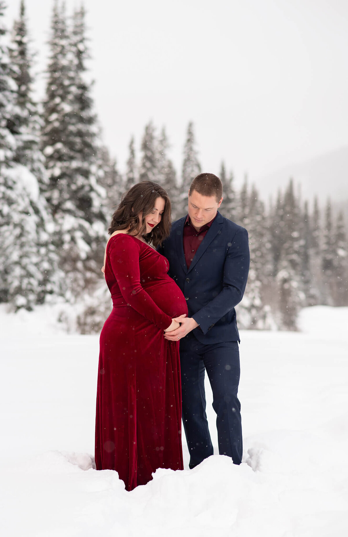 A couple is captured in Sun Peaks Kamloops. A husband and wife embrace against a stunning winter background. Woman is wearing a red velvet dress, husband in a suit.