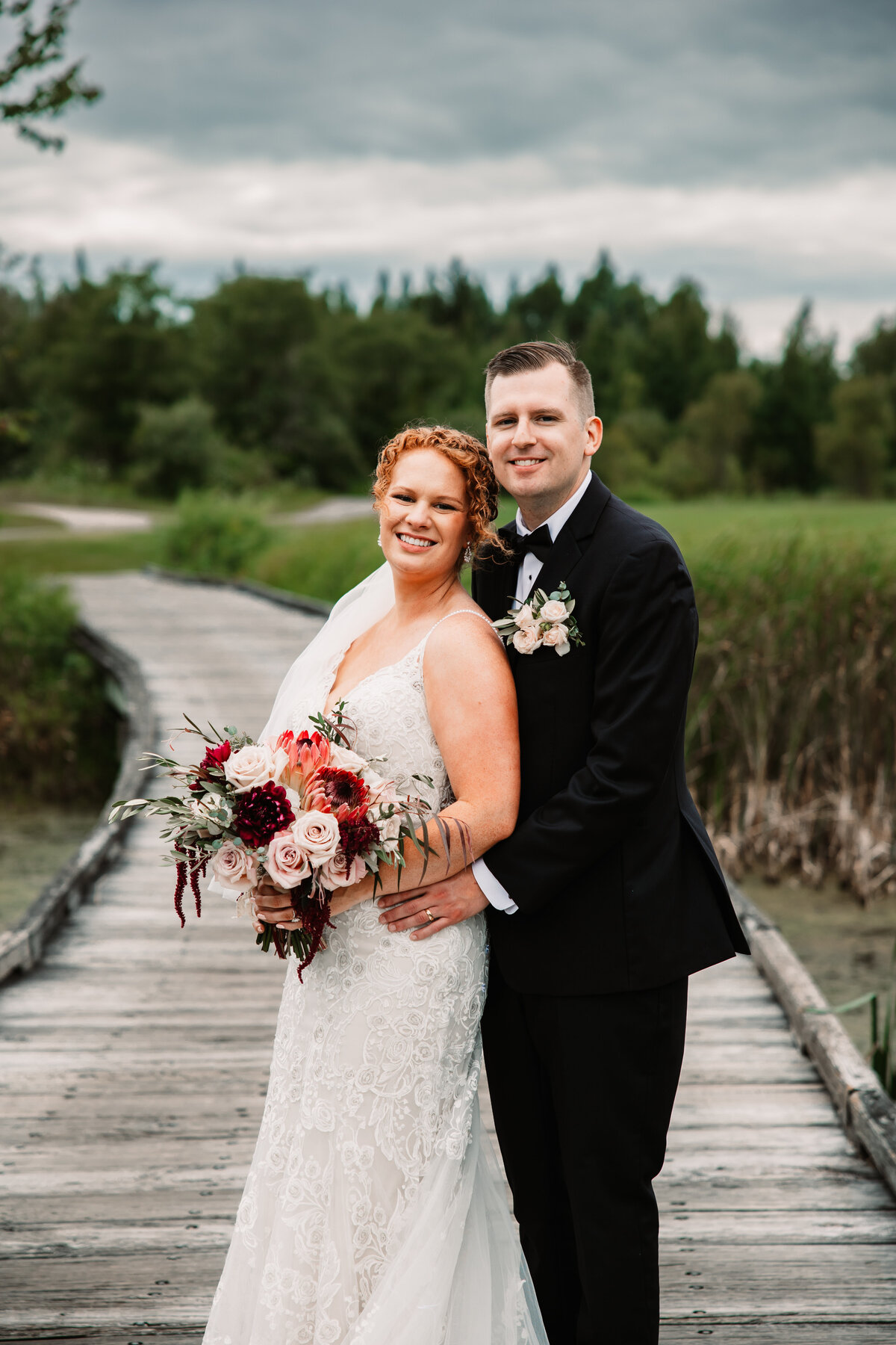 A bride and groom snuggle while standing on a wooden bridge over a pond
