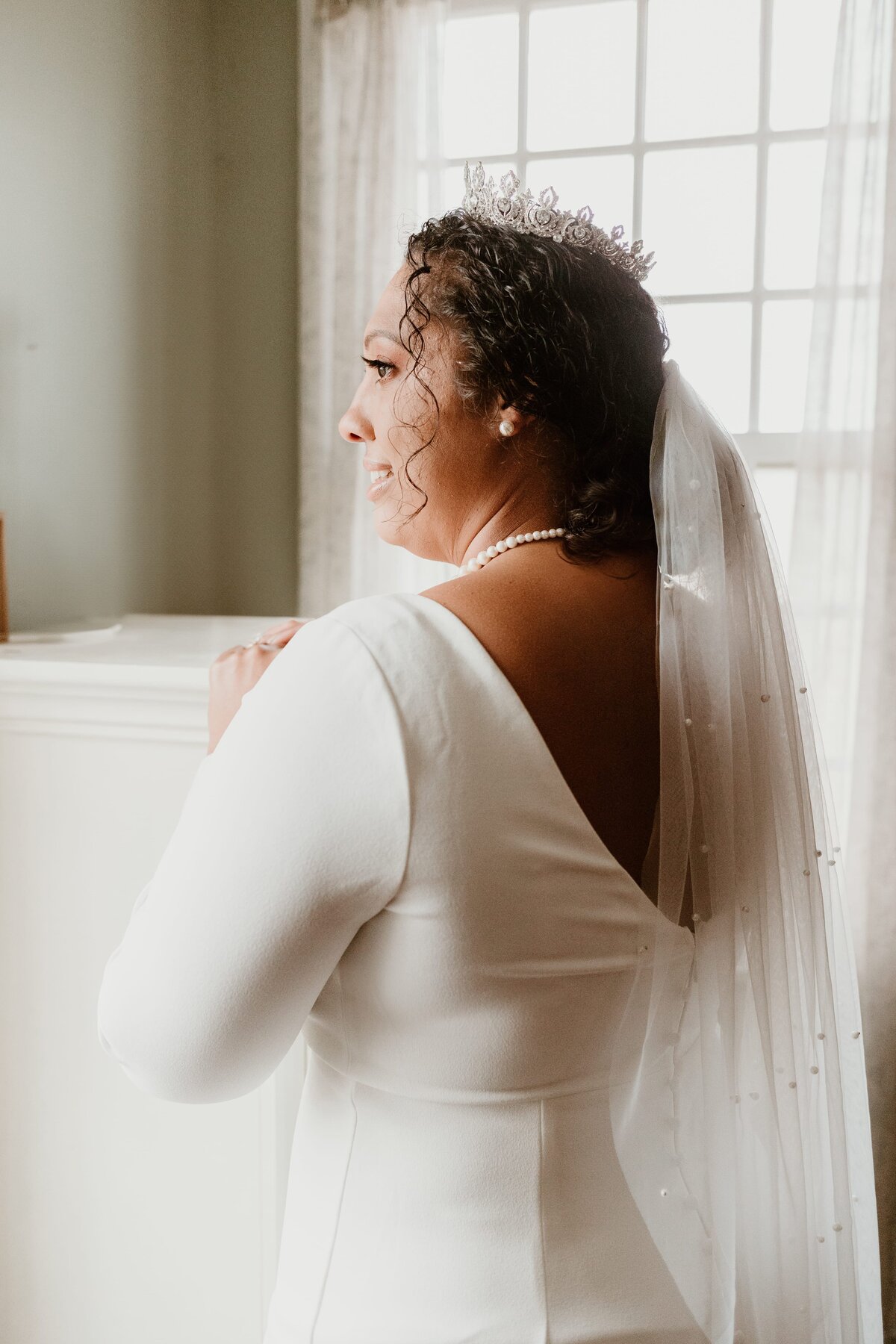 A radiant bride in a white gown and a bejeweled tiara gazes out a window, with soft light enveloping her from behind, creating a warm, ethereal atmosphere.