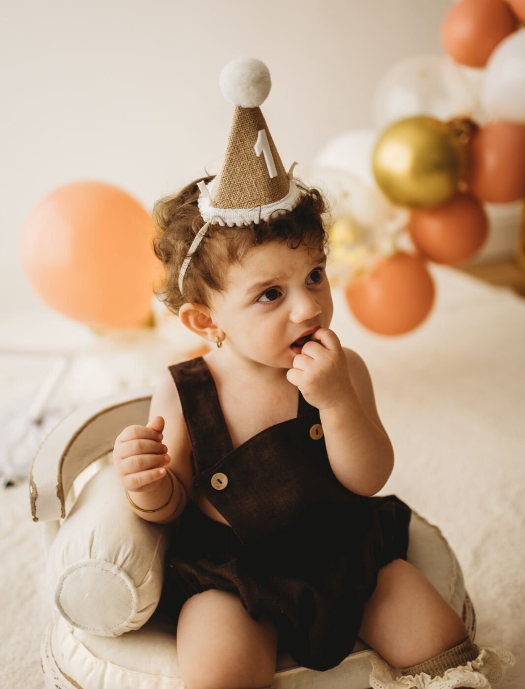 A one-year-old child wearing a burlap party hat and brown overalls sits with balloons behind them.