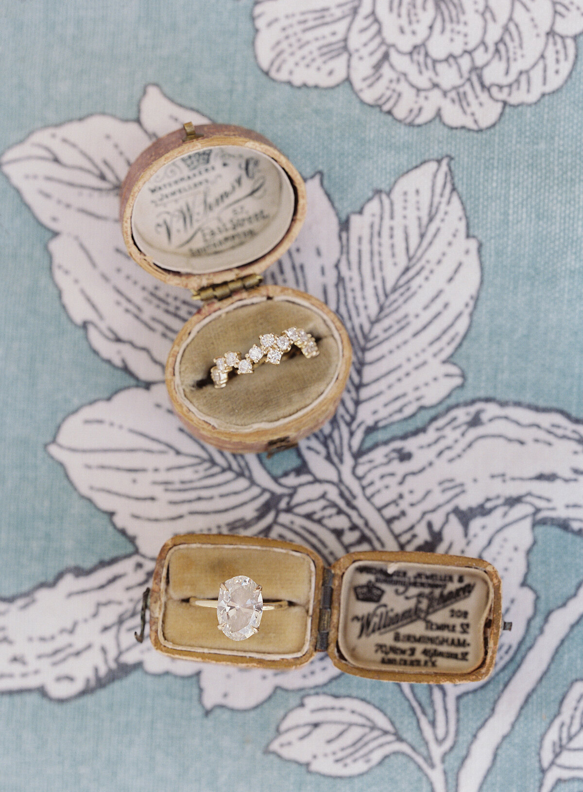 An antique ring box showcasing a stunning oval-cut diamond ring and a decorative diamond ring on a floral-patterned fabric background, complemented by a vintage jeweler's label. Perfect for Caribbean, Dominican Republic top tropical destination weddings with photographer Laura Gordon capturing the magic.