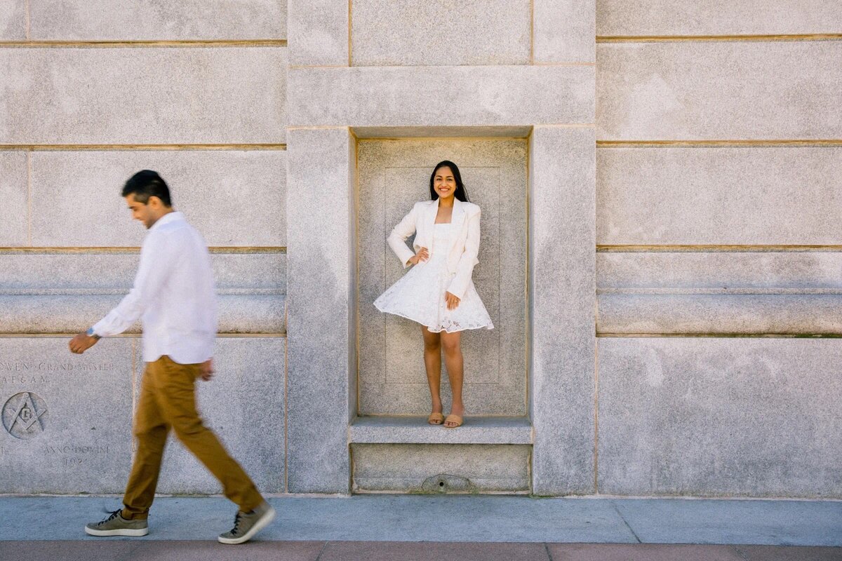 A woman in a white dress stands in an alcove while a man in a white shirt walks by, set against the backdrop of a stone building