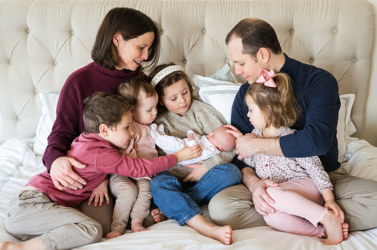 parents and siblings snuggling with newborn baby sister at home on the bed
