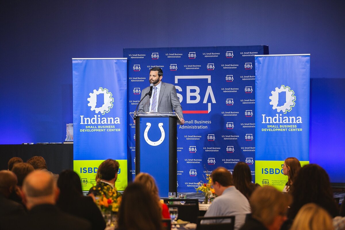 business-leader-speaking-at-small-business-administration-event-in-indianapolis Alt-Text: a-professional-speaker-in-a-suit-stands-at-the-podium-addressing-an-audience-at-a-small-business-administration-event-in-indianapolis-with-sba-and-indiana-small-business-development-center-banners-in-the-background