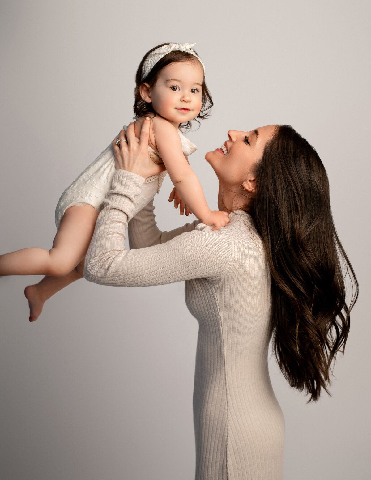 beautiful mother wearing cream dress holding her matching one year old baby up in the air while baby smiles at camera