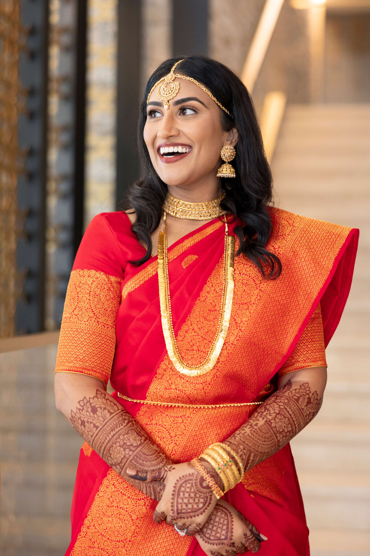 A woman in a red and orange saree stands smiling. She wears gold jewelry and has henna on her hands. The background features stairs and modern decor.
