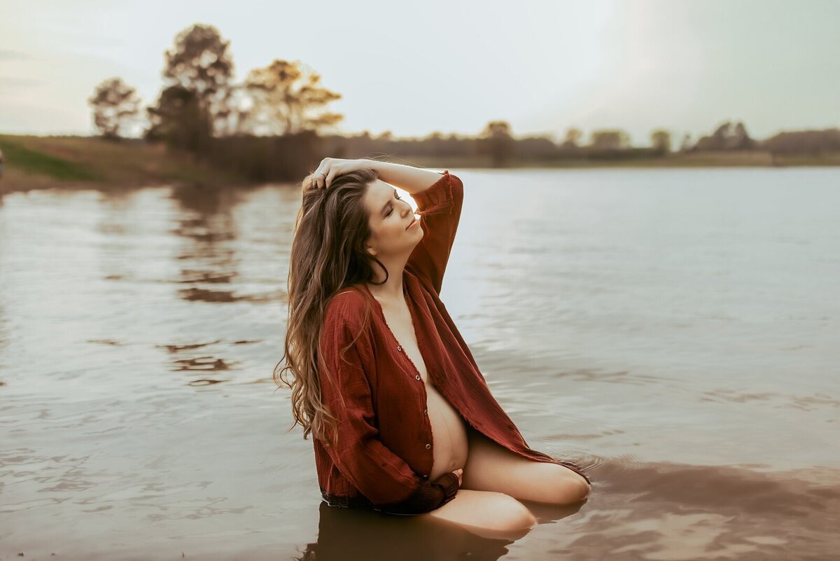 Pregnant woman sits in a lake, wearing a red shirt. She has one hand on her head and looks upward. Trees are in the background.