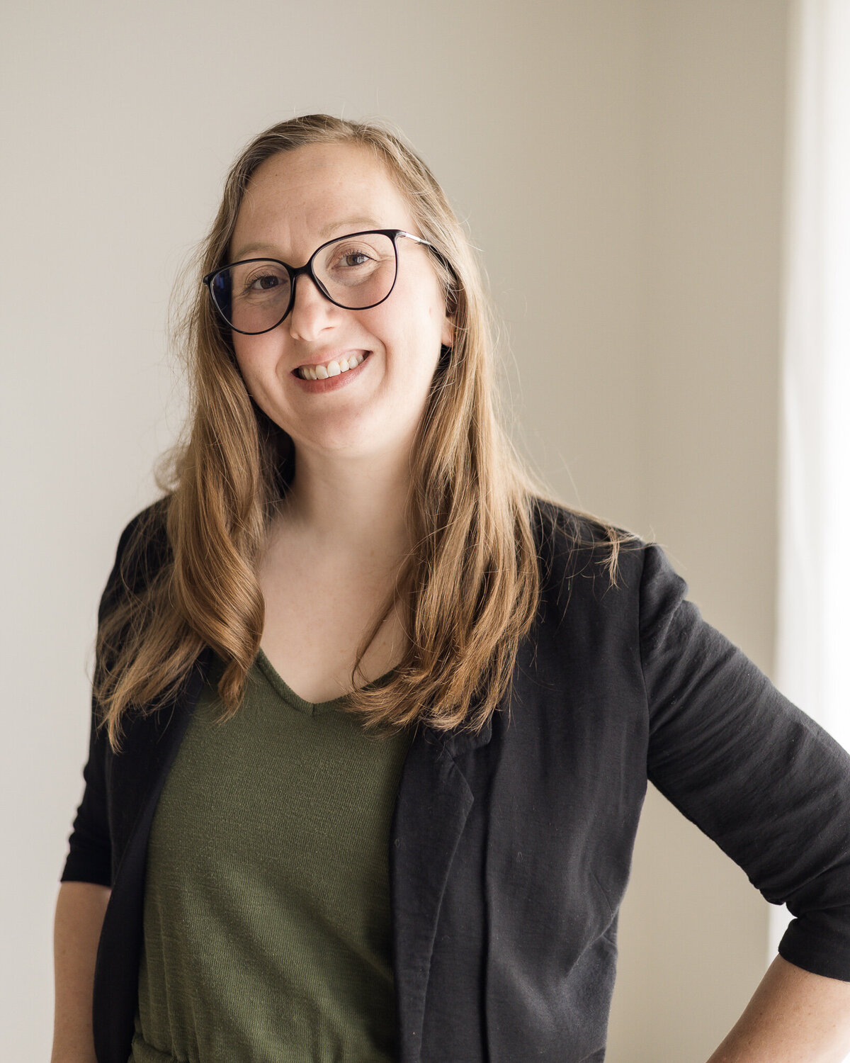 Strawberry blonde woman standing and slightly turned to the side, wearing glasses, a black dress jacket over a hunter green shirt and pants, wearing glasses, smiling at the camera