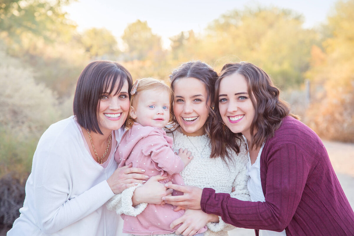 Portrait of three brown haired woman in white and maroon hugging and holding redheaded toddler girl