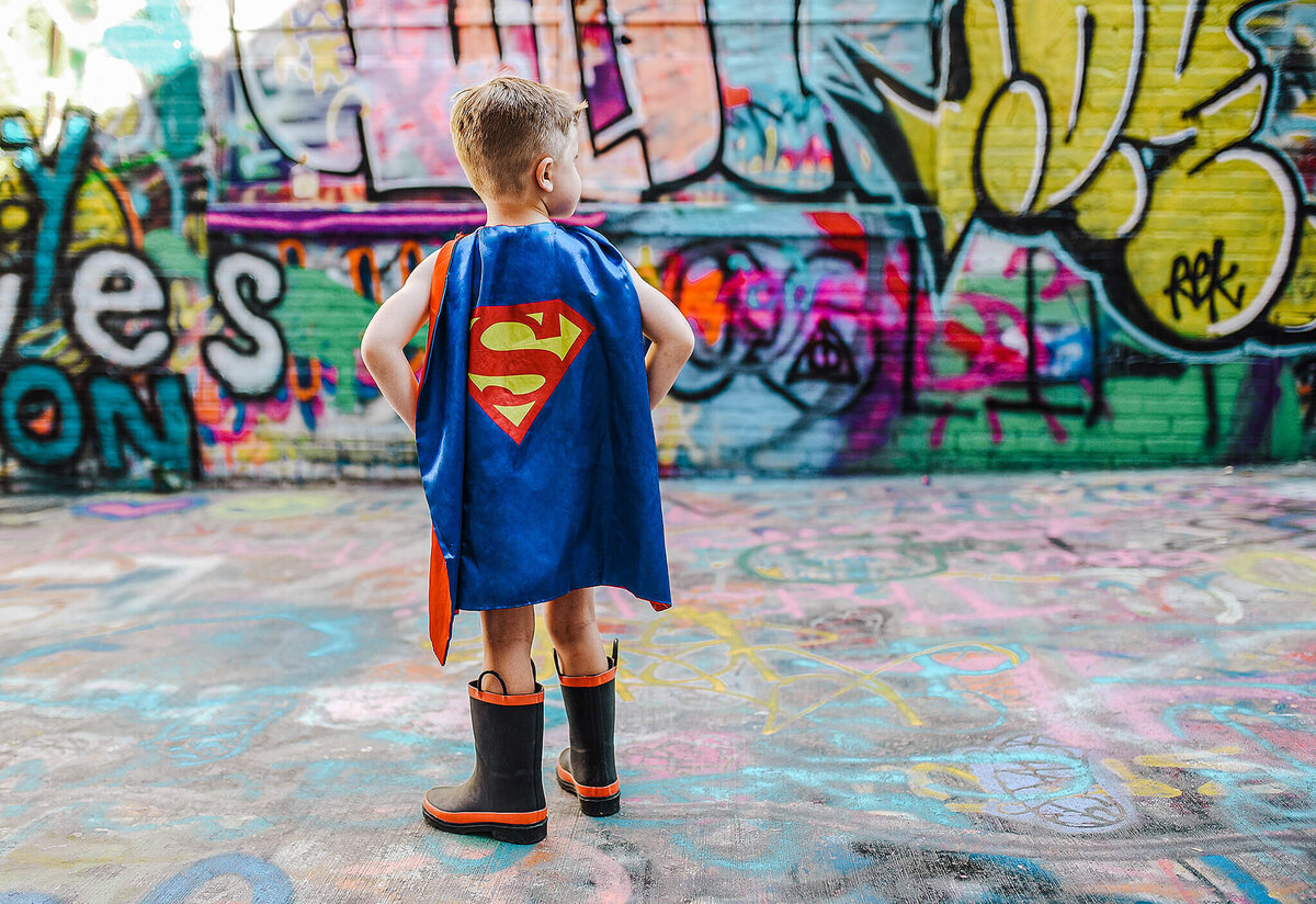 Little boy in black rain boots wearing a blue superman cape in graffiti alley near MICA in Baltimore Maryland