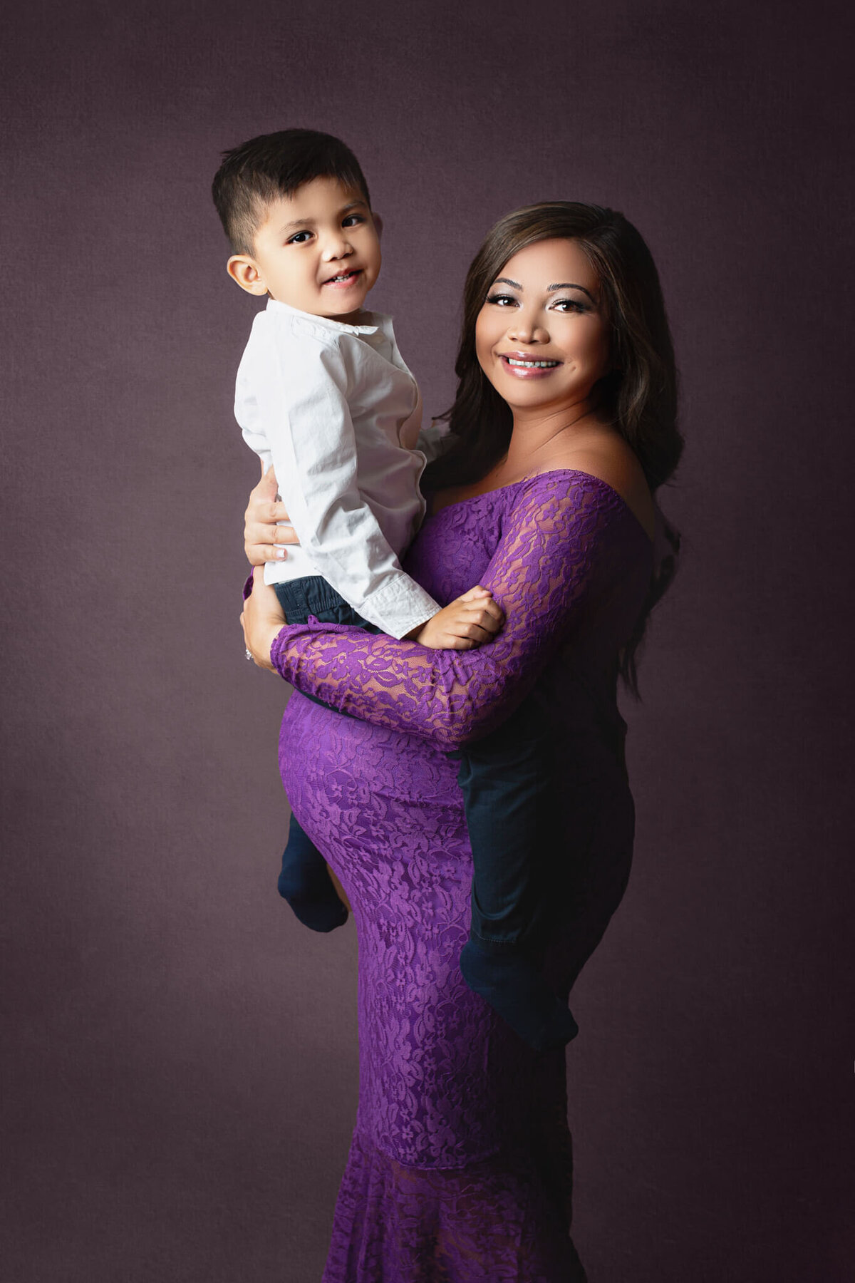 expecting mom holding her older son on top of her pregnant belly, wearing a purple lace maternity gown