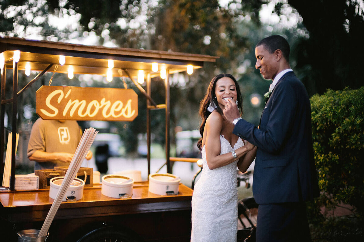 The bride and groom are eating in a S'mores station outdoors in Montage at Palmetto Bluff. Destination Image by Jenny Fu Studio