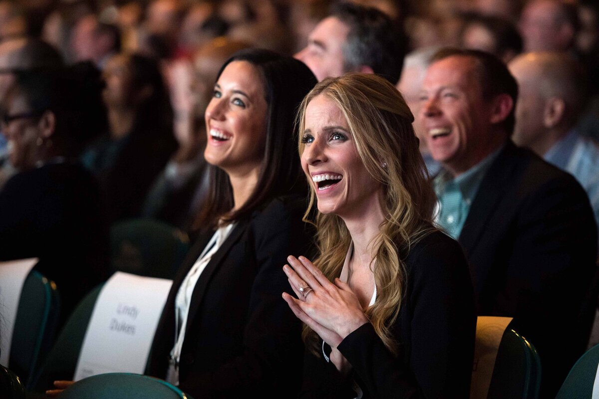 A crowd of people clap and smile in amusement while watching a speaker at their conference