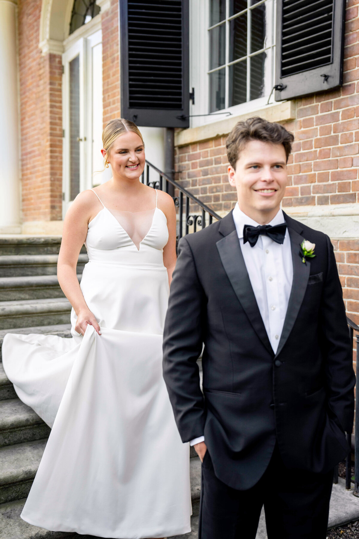 A bride in a white gown approaches a groom in a black tuxedo standing on stone steps. Both are outside a brick building with black shutters. The groom smiles as he looks away, while the bride holds the hem of her dress and smiles softly.