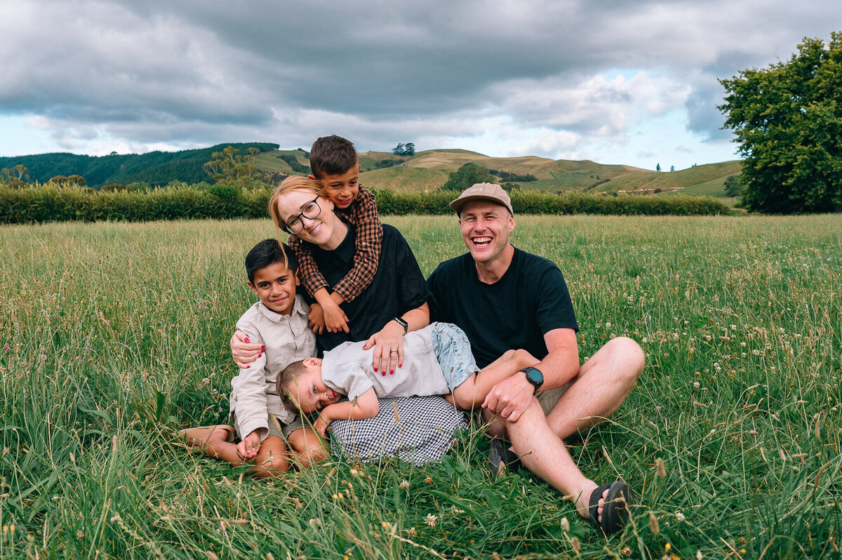family of 5 cuddling in a field of long green grasses with big grey clouds behind them