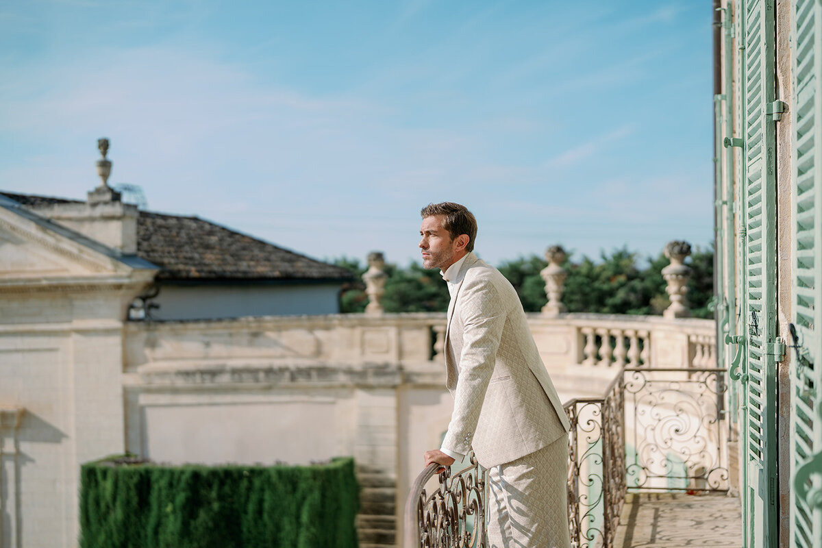 Groom standing on outdoor terrace overlooking gardens and stone architecture at Château de Tourreau in Provence.